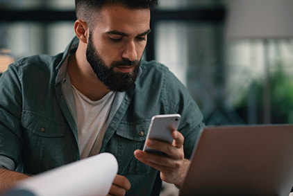 A man with a phone sitting in front of a laptop